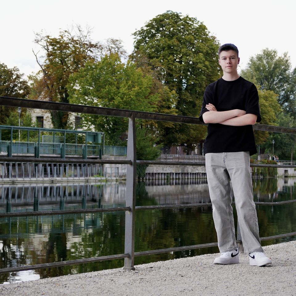 Tobi with his arms crossed at a railing by a river, trees and water behind him