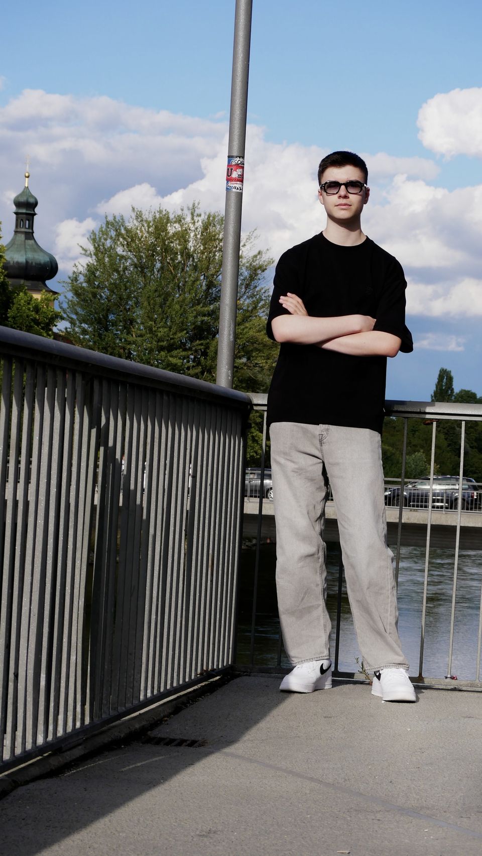 Tobi in sunglasses with his arms crossed on a bridge, a church tower and blue sky behind him