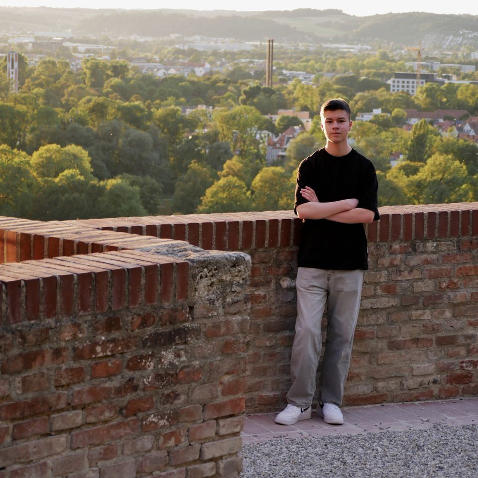 Tobi with his arms crossed at a brick parapet above the town, trees and hills behind him