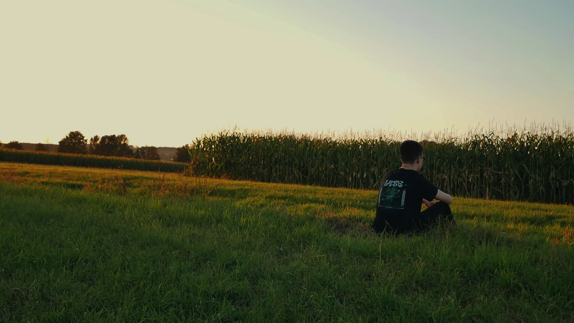 Camera shot from the music video: Tobi sitting in the grass in front of a corn field, filmed with a long lens